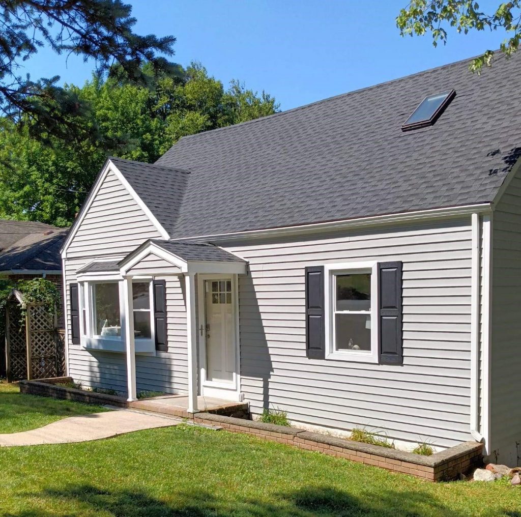 Small gray house with gable roof, skylight, black shutters, white door, lawn, and pathway.
