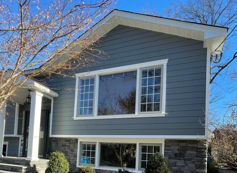 Two-story gray house with white trim, stone façade, large windows, clear blue sky.