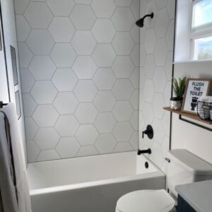 Modern bathroom with white hexagonal tiles, bathtub, shower, towel, shelf, plant, and toilet.
