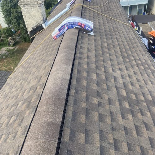 Rooftop with brown shingles, ridge vent, unopened shingle bundles, chimney, neighboring houses visible.