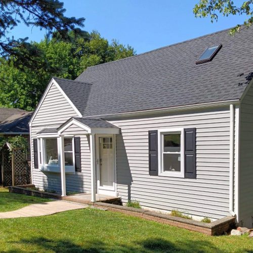 Small gray house with gable roof, skylight, black shutters, white door, lawn, and pathway.