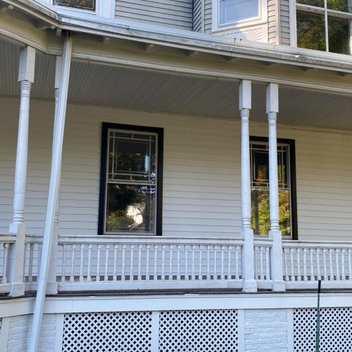 White two-story house with wraparound porch, decorative columns, large windows and black door.